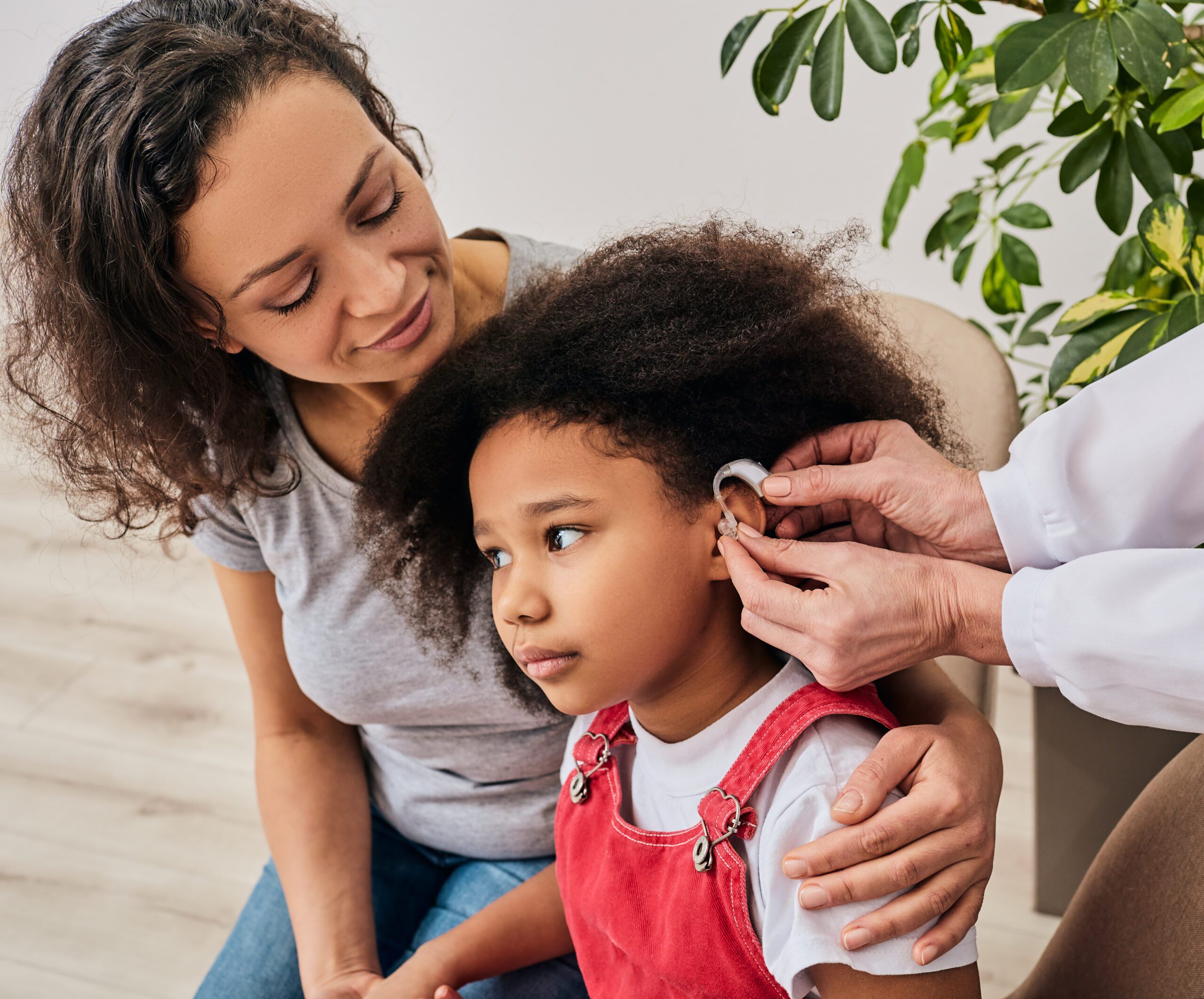A young child with curly hair sits next to an adult in an indoor setting. A person in a white coat is placing a hearing device in the child's ear.
